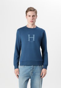 Felpa blu navy realizzata in tessuto morbido, con scollo rotondo e un grande logo "H" grigio chiaro con "Hackett" sottostante.