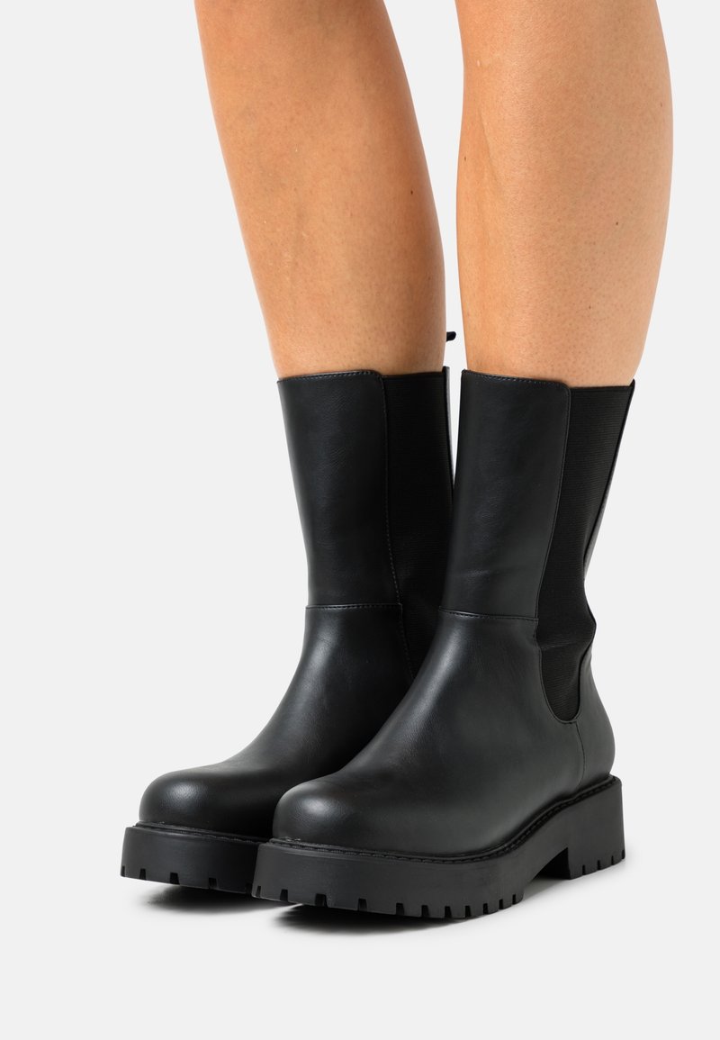 Bottes en cuir noires mi-mollet avec semelles épaisses, portées sur des jambes nues, sur un fond clair uni.