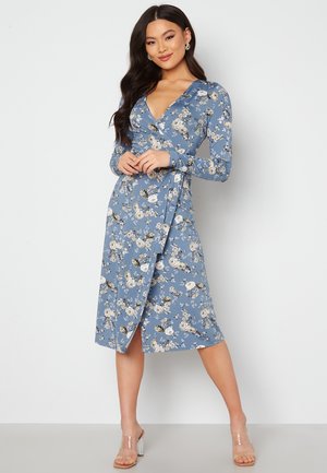 Blaues Blumen-Wickelkleid mit langen Ärmeln, V-Ausschnitt und seitlichem Schlitz; mit weißen und beigen Blumen auf glattem Stoff. Klare High Heels.