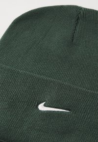 Zeleni pleteni beanie s rebrastom teksturom i bijelim Nike logotipom izvezenim sprijeda. Dizajn uključuje bešavni vrh i preklopni rub.