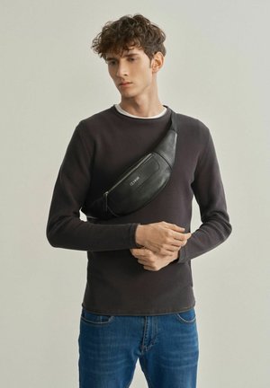 Czarna skórzana torba na pas z zamkiem błyskawicznym, noszona na ciele; model w ciemnym swetrze i niebieskich dżinsach, stojący na neutralnym tle.