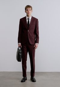 Βουργουνδί κοστούμι με στενή εφαρμογή, σακάκι με ένα κουμπί, λευκό πουκάμισο, μαύρη γραβάτα και μαύρη δερμάτινη τσάντα χειρός, σετ με ασορτί γυαλιστερά μαύρα παπούτσια.