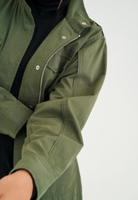 Olive green jacket with a zip front, snap fasteners, and a textured fabric. Features a chest pocket and a logo on the sleeve.