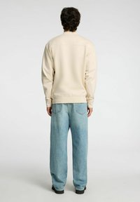 Hombre de pie de espaldas, vistiendo una camisa de manga larga beige, jeans de mezclilla azul claro y zapatos negros, frente a un fondo liso.