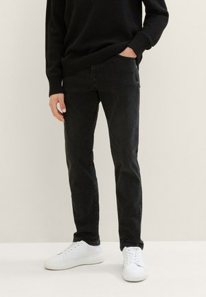 Osoba ubrana w czarne, dopasowane dżinsy, czarny sweter i białe tenisówki, stojąca z jedną ręką w kieszeni na jasnym tle.