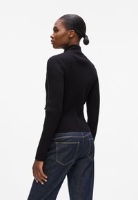 Maglione a collo alto nero a costine con maniche lunghe, dalla forma aderente e una texture liscia. Abbinato a jeans in denim scuro con cuciture gialle.