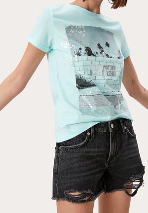 T-shirt en coton vert menthe avec un imprimé graphique, comprenant des accents argentés et une coupe décontractée, associé à un short en denim noir déchiré.