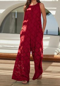 Jumpsuit rouge sans manches avec un motif en losanges texturé, jambes larges, poches latérales et une coupe décontractée. Fabriqué en tissu léger.