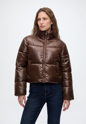 Une femme porte une doudoune matelassée marron brillante, à col montant et fermeture éclair sur le devant, assortie à un jean foncé, sur un fond uni.