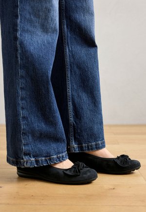 Bailarinas de ante negro con un detalle de lazo en la punta, combinadas con jeans de mezclilla de pierna ancha en color azul claro, sobre un suelo de madera.