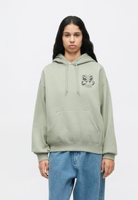 Fiatal személy, hosszú sötét hajjal, világoszöld kapucnis pulcsit visel pillangó mintával és laza kék farmernadrággal, fehér háttér előtt áll.