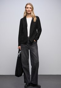 Svart texturerad kavaj med två knappar, i kombination med en vit topp och höga mörka jeans. Modellen håller en svart väska och bär svarta stövlar.