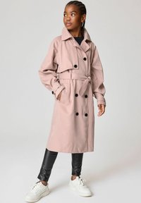 Ljusrosa trenchcoat med dubbelknäppta svarta knappar, bälte i midjan, rymliga fickor och uppvikta ärmar, matchad med svarta leggings och vita sneakers.
