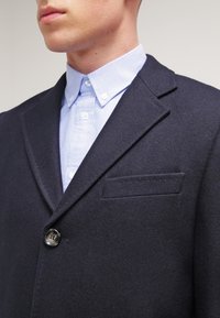 Marinblå ullblazer med spetsig slag, enkel knappstängning och bröstficka. Bärs över en ljusblå skjorta med knappar.