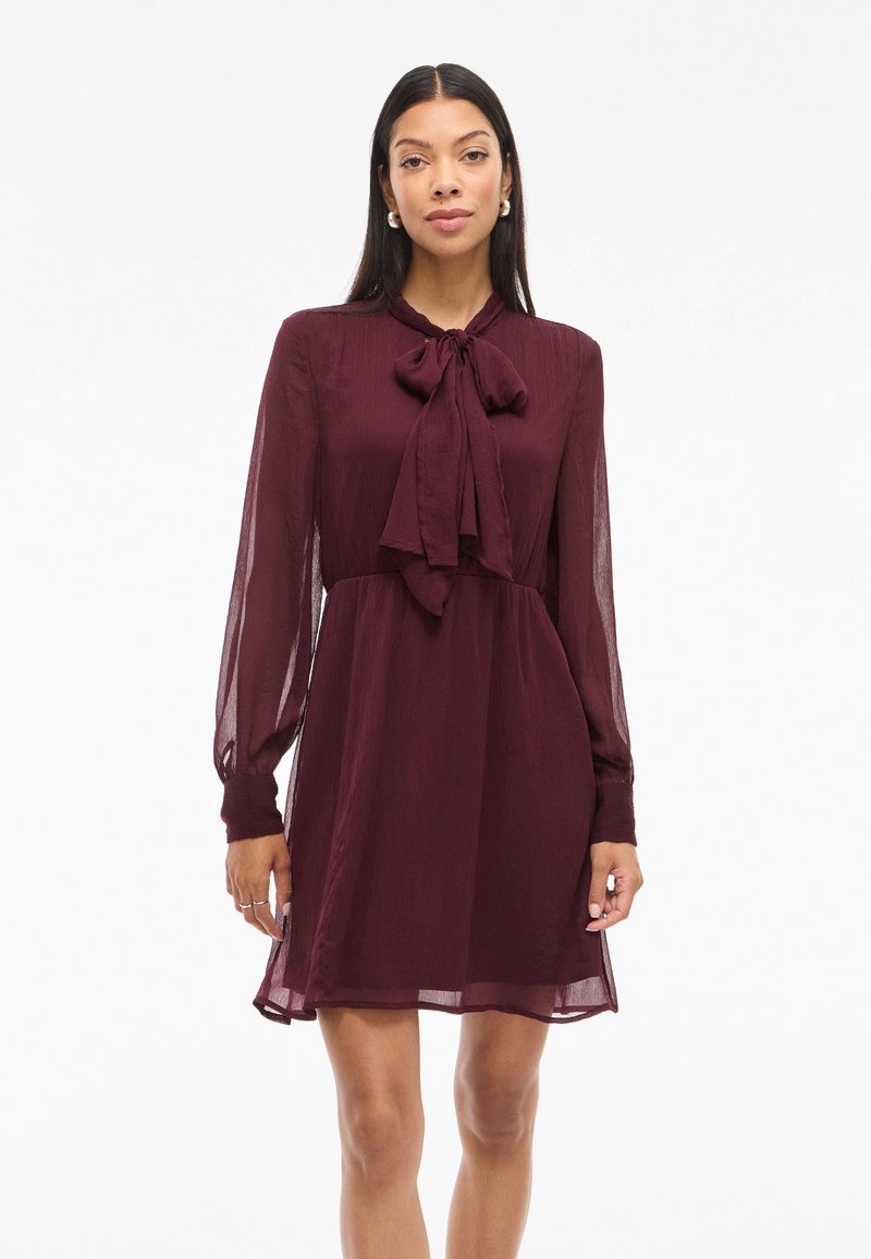 Kobieta stojąca w burgundowej sukience z długim rękawem, z wiązanym krawatem przy szyi, przezroczystymi rękawami i lekko rozszerzaną spódnicą.