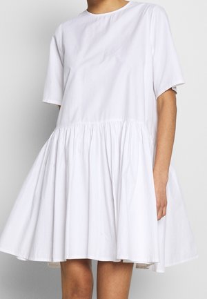 Løs, hvid kjole med korte ærmer, samlet talje og udvidet skørt, båret af en person mod en ensfarvet baggrund.