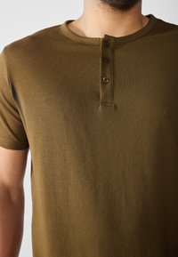 Tricou scurt cu mâneci scurte de culoare verde măsliniu, fabricat din bumbac moale, având un gât rotund și o deschidere cu trei nasturi în față. Textură netedă.