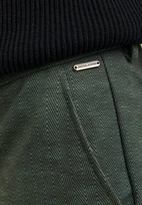 Lähikuva vihreistä herringbone-kuosisista housuista, joissa on metallinen Jack & Jones -logotagi mustan ribbireunuksen alla.