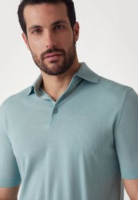 Gaiši zilas polo krekls, izgatavots no mīkstas auduma, ar apkakles izgriezumu, īsām piedurknēm un divu pogu aizdari. Gluda tekstūra.