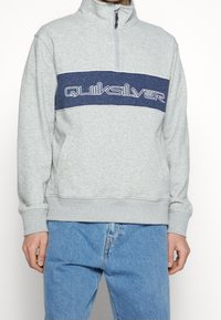 Persona vestida con sudadera gris claro de media cremallera y franja azul marino, con el texto "Quiksilver" en el pecho, combinada con jeans azules.