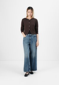 Vrouw staat in een donkerbruine gebuttonde cardigan, wijdvallende blauwe jeans en zwarte flatjes tegen een effen witte achtergrond.