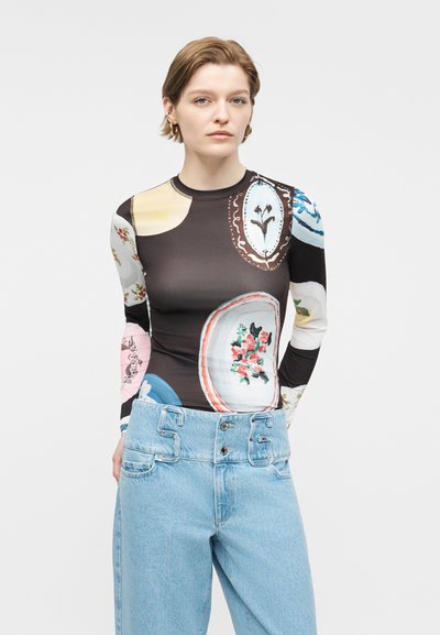 Νεαρή γυναίκα που φοράει μακρυμάνικο τοπ με πολύχρωμα αφηρημένα λουλουδένια μοτίβα και ψηλόμεσα ανοιχτόχρωμα τζιν τζιν παντελόνια μπροστά σε απλό φόντο.