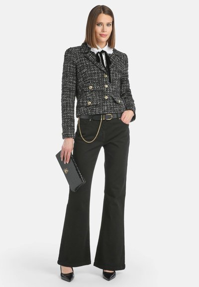 Mustavalkoinen tweed-takki, jossa on kaksiriviset kultanapit, yhdistettynä leveneviin mustiin housuihin. Musta clutch-laukku ja korkokengät täydentävät asun.