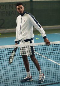 Weiße Reißverschlussjacke mit marineblauen Akzenten, weiße Shorts und marineblaue Sportschuhe. Steht neben einem Tennisnetz auf einem blauen Platz.