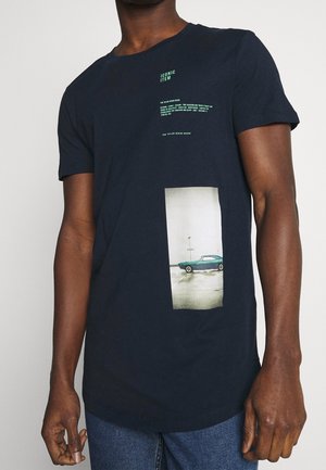Homme portant un T-shirt bleu marine avec un petit texte vert "ARTICLE ICONIQUE" et une photo d'une voiture vintage verte garée sur un pavé mouillé.