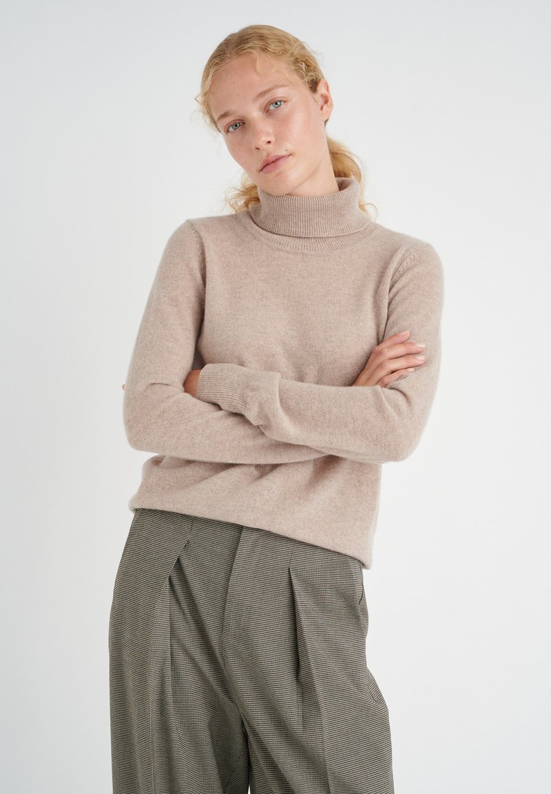 Beige, knitted turtleneck sweater with long sleeves; soft texture. Worn with high-waisted, houndstooth patterned trousers.