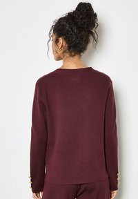 Suéter burdeos con un corte holgado, presenta un escote ancho y acentos de botones dorados en los puños, fabricado con una suave tela texturizada.