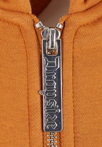 Sweat-shirt zippé orange avec un tissu texturé et un tirette de fermeture éclair en métal brillant portant le mot "Dropout" en lettres en gras et stylisées.