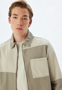 Beige en taupe tweekleurige jas met een ritssluiting, voorzien van een borstzak en een kraag. Gemaakt van een geweven katoenmix.