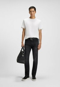 T-shirt bianco a maniche corte, jeans neri, sneakers nere con suola bianca, che tiene in mano una borsa nera con trama, in piedi contro uno sfondo neutro.