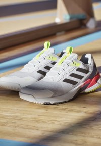 Białe buty sportowe z czarnymi paskami, szary górny materiał z siateczki, różowe i limonkowe akcenty, teksturowana podeszwa oraz wzmocniony obszar palców.