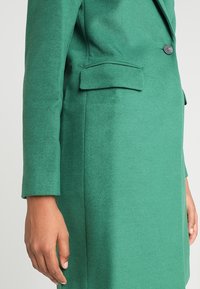 Blazer verde de tela texturizada, con un botón único, bolsillo con solapa lateral y mangas largas entalladas.