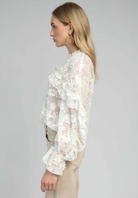 Blouse blanche avec des manches volantées et un motif floral texturé, dotée d'un col montant et d'un tissu doux et transparent.