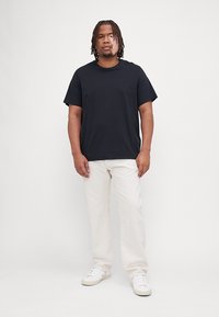 Camiseta negra de manga corta en tejido suave, combinada con pantalones rectos blancos y zapatillas blancas, de pie contra un fondo neutro.