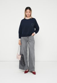 Felpa blu navy con design a cuore, vestibilità oversize, abbinata a jeans grigi a gamba larga, una borsa rosa e scarpe rosse con fiocco.
