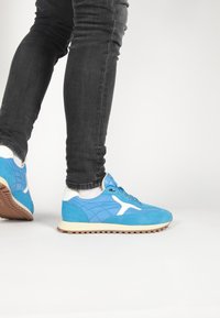 Blue suede sneaker with white accents, textured upper, rubber sole, and lace closure, paired with black slim-fit jeans.