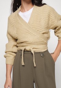 Femme portant un pull beige en maille cache-cœur noué à la taille, sur un haut blanc, et un pantalon taille haute vert olive avec des boutons en détail.