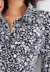 Námořnická modrá květinová košile s bílými botanickými vzory, zapínání na knoflíky a klasickým límcem z texturované látky. Červený akcent uvnitř límce.