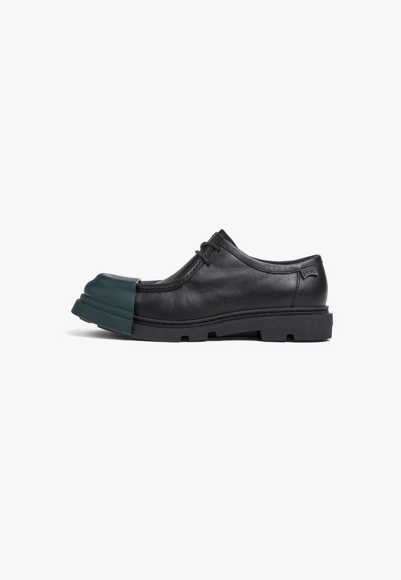 Chaussure en cuir noir avec un bout en caoutchouc vert distinctif. Présente un design à profil bas, un système de laçage et une semelle robuste pour une meilleure adhérence.