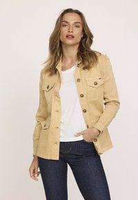 Veste en coton beige avec des boutons, deux poches poitrine et une coupe décontractée. Portée par-dessus une chemise blanche et un jean foncé. Texture lisse.