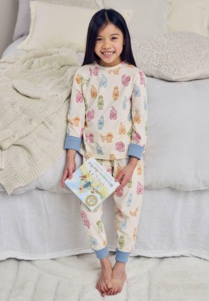 Smiling girl in colorful dog-print pajamas holding a children's book, standing barefoot next to a bed with textured blankets and pillows.