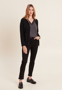 Schwarze Zip-Up-Jacke, graues Top mit Spitzenbesatz, schwarze eng anliegende Hose und glänzende schwarze Slipper. Glatte Textur, maßgeschneiderte Passform und umgeschlagene Ärmel.