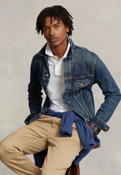 Jeune homme avec des dreadlocks portant un polo blanc, une veste en jean, un pantalon beige et un pull bleu noué autour de la taille, assis sur un tabouret.