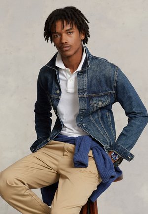 Giovane uomo con dreadlocks che indossa una polo bianca, una giacca di jeans, pantaloni beige e un maglione blu annodato intorno alla vita, seduto su uno sgabello.