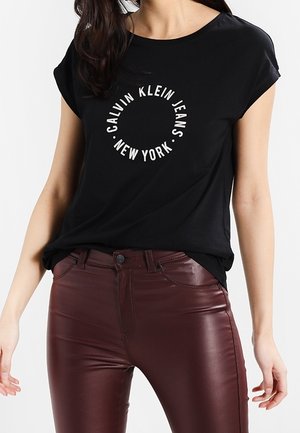 Femme portant un t-shirt noir Calvin Klein Jeans avec un logo blanc circulaire et un pantalon en cuir bordeaux foncé taille haute, debout devant un fond blanc.