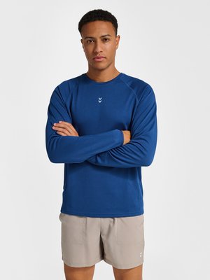 Mann trägt ein blaues langärmeliges Sportoberteil und beige Shorts, steht mit verschränkten Armen vor einem schlichten weißen Hintergrund.